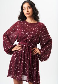 Burgundy dress with long puff sleeves, belted waist, and star-like metallic patterns. Sheer fabric adds texture and lightness.