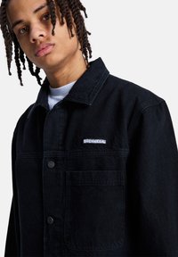 Νεαρός με πλεγμένα μαλλιά, φοράει μαύρο τζάκετ με κουμπιά, με μπροστινή τσέπη και λευκό λογότυπο DC Shoes στο στήθος.