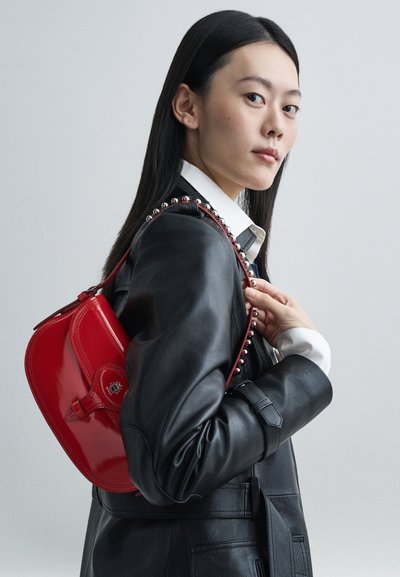 Röd handväska i lackläder med rundad form, med en dekorativ rem med silvernitar, fäst med en låtdetalj.