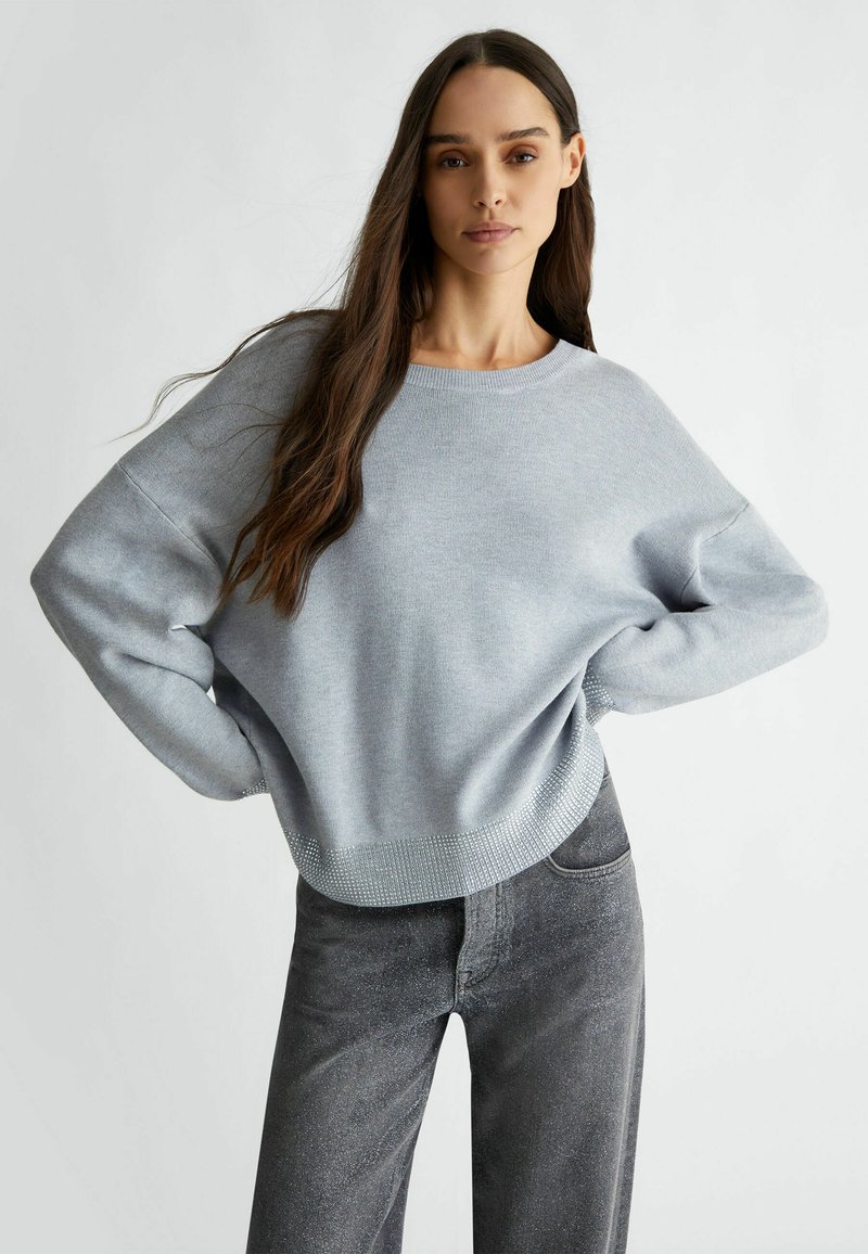 Maglione grigio chiaro con scollatura rotonda, spalle scese e orlo testurizzato. Abbinato a jeans grigi scuri a vita alta.
