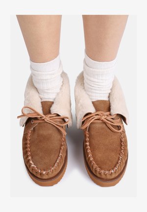 Brązowe zamszowe kapcie z miękkim beżowym futrem w środku, związane jasnobrązowym sznurkiem, z teksturowaną podeszwą i dekoracyjnym przeszyciem.