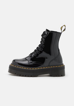 Bottes de cheville en cuir verni noir avec une semelle plateforme épaisse, orteil rond et couture jaune. Elles disposent de lacets et d'une languette à l'arrière.