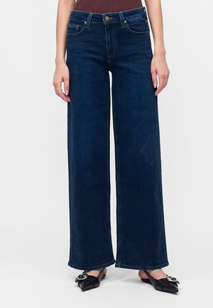 Femme portant un jean bleu foncé à jambes larges et des ballerines noires pointues avec une boucle argentée sur fond uni.