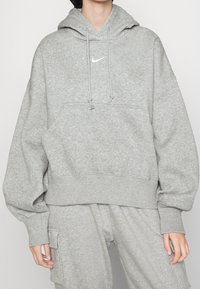 Osoba oblečená v světle šedé mikině Nike a odpovídajících teplácích se boční kapsou, stojící na neutrálním pozadí.