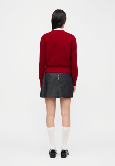 Woman standing facing away wearing a red sweater, dark gray skirt, white knee-high socks, and black shoes against a plain background.