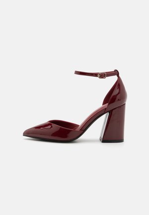 Burgundy patent leather high-heeled pump with a pointed toe, ankle strap, and chunky heel. Smooth texture and sleek design.