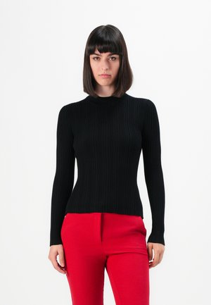 Vrouw met steil donker haar en een pony, gekleed in een zwart geribbeld langemouwen top en felrode broek, tegen een witte achtergrond.