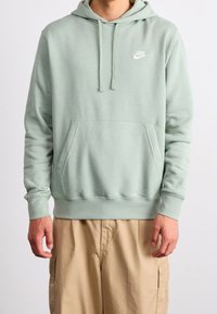 Persona vistiendo una sudadera Nike verde claro con bolsillo frontal y cordones, combinada con pantalones cargo beige, de pie frente a un fondo liso.