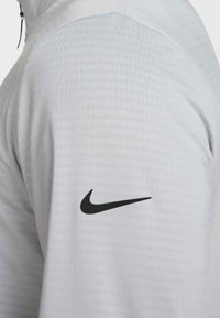 Nike világosszürke sportpulóver sima textúrával, félcipzáras kialakítással és egy hangsúlyos fekete Swoosh logóval a bal vállon.