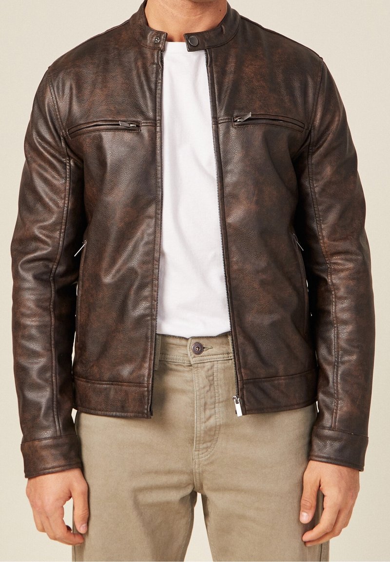 Man draagt een bruin leren jack over een wit t-shirt en beige broek, met volledig uitgestrekte mouwen van het jack en een gedeeltelijk openstaande rits aan de voorkant.