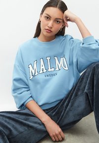 Világoskék pulóver "MALMÖ" fehér hímzett logóval, sötét kordbársony nadrággal párosítva, laza szabással és bordázott mandzsettákkal.