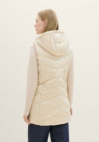 Beige Steppweste mit Chevron-Steppung, ausgestattet mit einer Kapuze und aus leichtem Material. Das Model trägt darunter einen hellen Pullover.