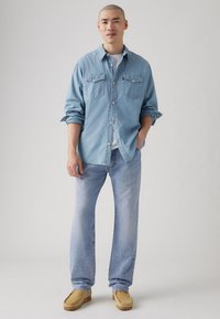 Lyseblå denimskjorte med to brystlommer, brukt over en hvit t-skjorte, kombinert med lyseblå jeans og beige slip-ons.