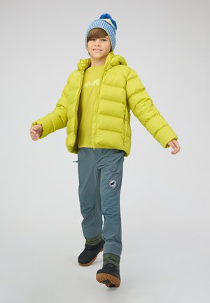 Jongen loopt, draagt een groene donsjas, een blauwe gebreide muts, grijze broek en zwarte laarzen op een effen lichte achtergrond.