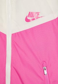 Rožinė lengva striukė su baltais akcentais, su priekiu užtrauktuku ir rožiniu "Nike" logotipu viršutiniame kairiajame kampe. Lygus audinys.