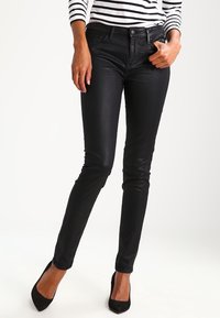 Svarta tajta jeans i ett mjukt, stretchigt material med framfickor, midja med bältesöglor och en subtil glans. Bärs med svarta klackar.