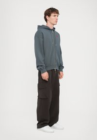 Un sweat à capuche gris zippé avec une poche avant et un logo rouge, associé à un pantalon cargo noir ample avec des poches latérales et des chaussures blanches.