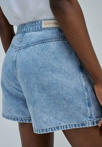 Pantalones cortos de mezclilla azul claro con cintura ajustada, que presentan costuras visibles y una etiqueta de marca. La tela muestra una ligera textura desgastada.