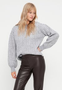 Pull en maille gris clair avec une coupe décontractée, motif en losanges texturé et manches ballon, associé à un pantalon en cuir marron ajusté.