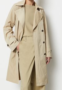 Trench coat beige con un design doppiopetto, colletto ampio e dettagli in bottone, abbinato a un maglione chiaro e pantaloni coordinati.