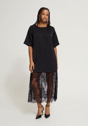 Femme aux cheveux tressés portant une robe noire à manches courtes avec un jupon en dentelle transparente et des chaussures à talons hauts avec bride à la cheville noires.