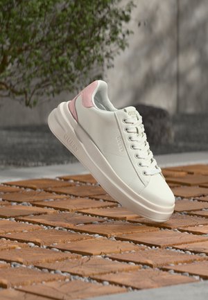 Λευκό sneaker χαμηλού προφίλ με ροζ λεπτομέρεια στη φτέρνα, κορδόνια μπροστά και παχύ πέλμα, τοποθετημένο πάνω από πλακάκια σε εξωτερικό χώρο.