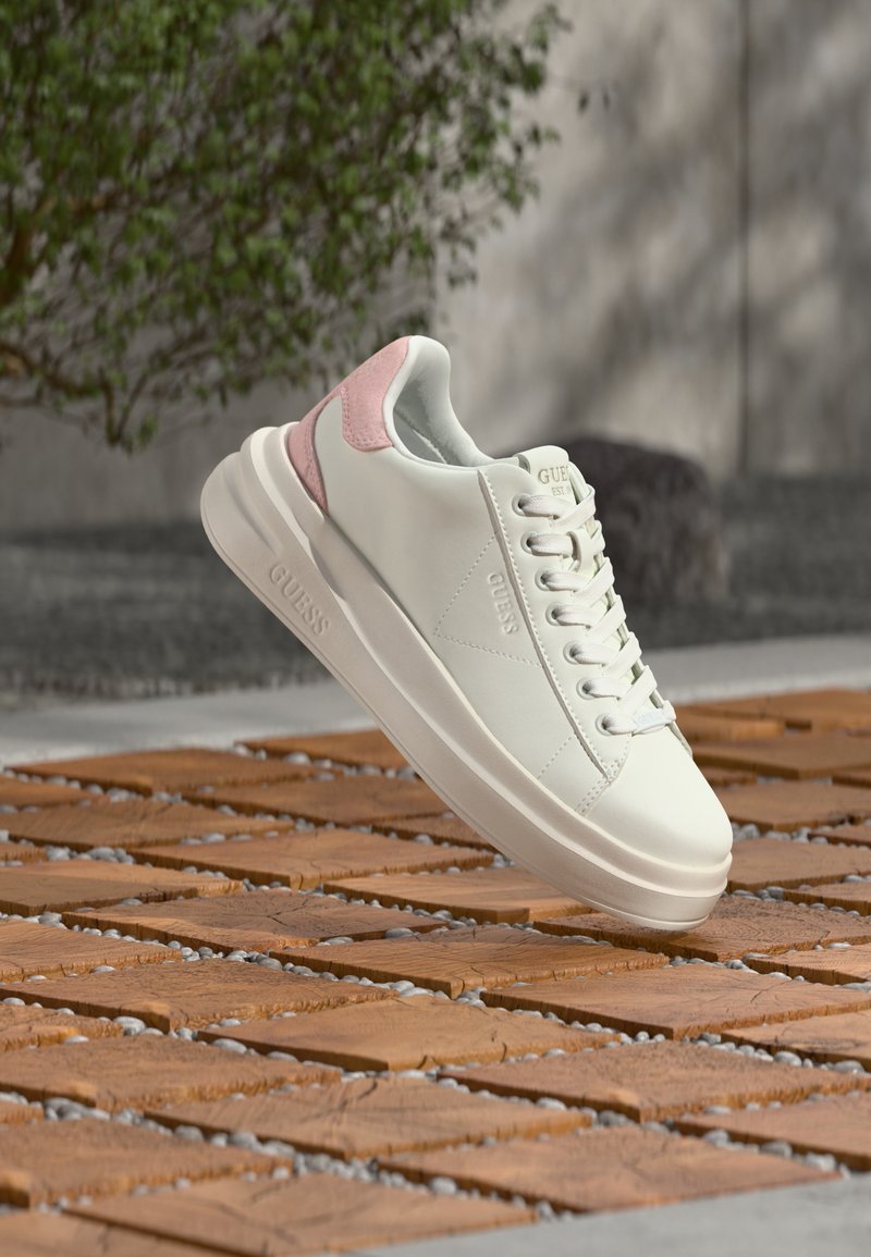 Λευκό sneaker χαμηλού προφίλ με ροζ λεπτομέρεια στη φτέρνα, κορδόνια μπροστά και παχύ πέλμα, τοποθετημένο πάνω από πλακάκια σε εξωτερικό χώρο.