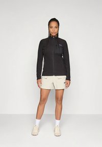 Μαύρο φλις μπουφάν με φερμουάρ και υφή στην επιφάνεια, συνδυασμένο με ανοιχτό μπεζ σορτς που διαθέτει δύο μπροστινές τσέπες. Αθλητικά παπούτσια και κάλτσες σε ουδέτερους τόνους.