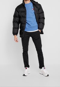 Giubbotto puffo nero con finitura lucida, felpa blu con logo, pantaloni neri e sneakers bianche con dettagli neri.