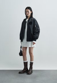 Veste bomber noire à manches oversize, poche zippée sur le bras, portée sur une robe blanche, associée à des bottes hautes marron et un petit sac noir.