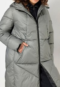 Cappotto imbottito grigio chiaro con cappuccio, dotato di chiusura con zip, tasche laterali e finitura testurizzata, esposto con un motivo di cuciture a triangolo.