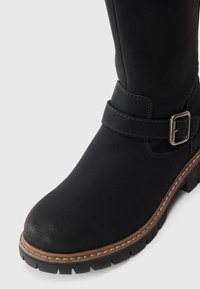Schwarze Knöchelstiefel mit glatter Textur, ausgestattet mit einem silbernen Schnallenriemen und einer kontrastierenden braunen Sohle, die durch weiße Stichdetails hervorgehoben wird.