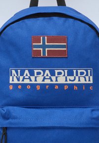 Sac à dos en tissu bleu avec un patch marron arborrant un drapeau norvégien, et un logo blanc indiquant "NAPAPIJRI" avec des accents orange en dessous.
