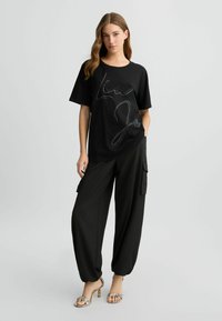 Zwarte kortekraag T-shirt met een wit cursief logo, gecombineerd met losse zwarte cargobroeken en slangenprint hakken. Glad stoffen textuur.
