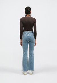 Top marrón ajustado de manga larga combinado con jeans acampanados azul claro. Se observa un detalle de dobladillo desgastado y dos bolsillos traseros. Zapatillas blancas completan el look.