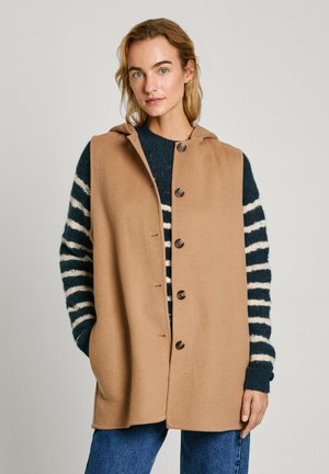 Beige sleeveless coat with a hood, featuring large brown buttons and a straight cut. Underneath, a navy striped sweater is visible.