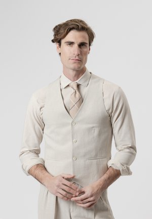 Hombre con cabello castaño claro, vestido con camisa beige, chaleco a juego y corbata a rayas, de pie con las manos entrelazadas al frente sobre un fondo blanco.