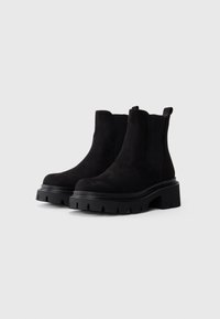 Une paire de bottes Chelsea en suède noir avec des semelles en caoutchouc épaisses et des panneaux latéraux élastiques sur un fond blanc.
