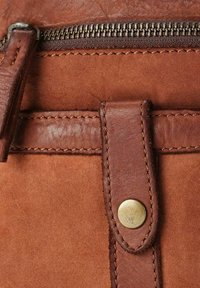 Dettaglio di una borsa in pelle marrone con zip, una tracolla in pelle con borchia in ottone e cuciture a contrasto su una superficie testurizzata.