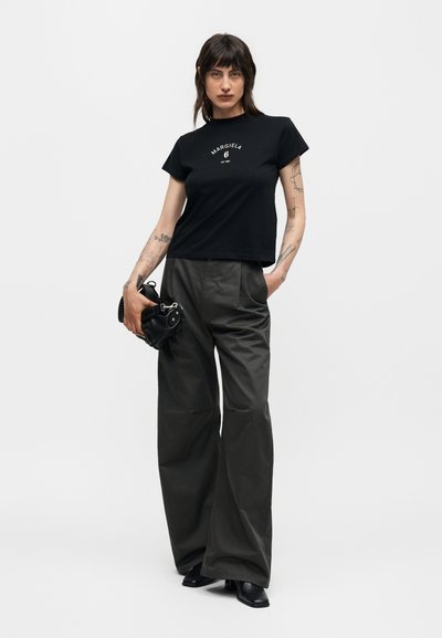 Γυναίκα με τατουάζ φοράει μαύρο t-shirt Margiela, φαρδιά μαύρη παντελόνα, μαύρες μπότες και κρατάει μια μικρή μαύρη τσάντα.