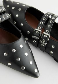 Ballerine nere in cuoio con punta appuntita, arricchite da borchie circolari argentate e doppi cinturini con fibbia, caratterizzate da una superficie liscia e una fodera interna marrone.