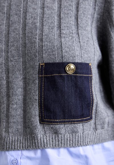 Suéter gris de canalé con un bolsillo de mezclilla oscura en contraste, que presenta un detalle de botón dorado. La textura de la tela es suave y de punto.