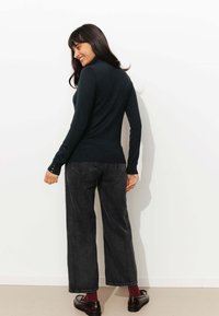 Pull à col roulé bleu marine avec des manches longues, poignets contrastés avec des boutons. Porté avec un jean large gris foncé et des chaussures noires.