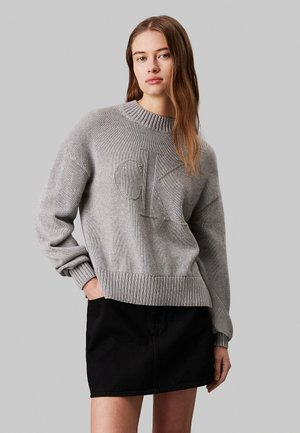 Light grey knit sweater with ribbed neckline and cuffs, featuring an embossed "CK" logo on the front. Worn over a black skirt.