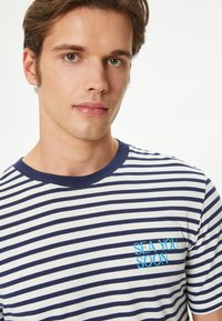 Camiseta de algodón a rayas con rayas horizontales en azul marino y blanco, escote redondo y texto turquesa que dice "TE VEO PRONTO" en el pecho.