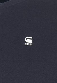 Marinblå t-shirt med ribbad struktur, med en vit logotyp som lyder "RAW" på vänster sida av bröstet.