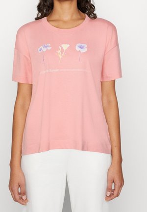 Nainen, joka pukeutunut väljään vaaleanpunaiseen T-paitaan, jossa on kolme pastelliväristä kukkakuvaa ja valkoinen teksti, yhdistettynä valkoisiin housuihin, yksiväristä taustaa vasten.