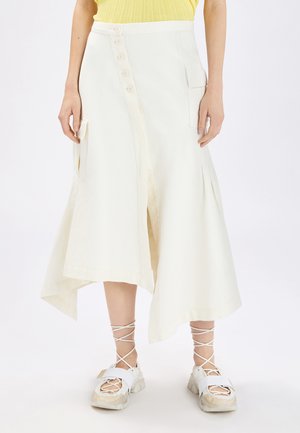 Donna che indossa una gonna midi asimmetrica colore bianco sporco con bottoni e tasche con patta, abbinata a sneakers bianche con lacci e una maglietta gialla.