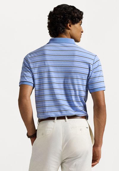 Άνδρας με σγουρά μαλλιά, φορώντας ένα ανοιχτό μπλε πόλο με λευκές και μαύρες ρίγες, ανοιγμένο μέσα σε λευκά παντελόνια με καφέ ζώνη.