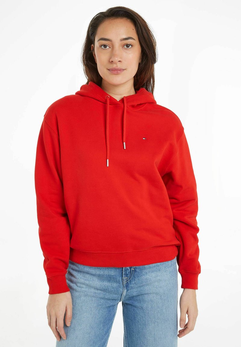 Sweatshirt à capuche rouge avec une coupe décontractée, des poignets et un ourlet côtelés, présentant un petit logo sur la poitrine. Associé à un jean bleu clair.